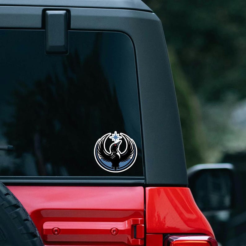May include: A circular sticker featuring a stylized black bird with outstretched wings, a blue and white border, and a white starburst design. The sticker is affixed to the rear window of a red vehicle.