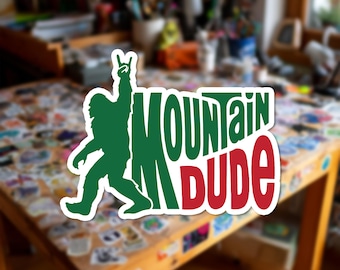 Può includere: Un adesivo fustellato raffigurante la sagoma verde di una figura di Bigfoot che fa un gesto rock-and-roll con la mano accanto al testo "MOUNTAIN DUDE" in grandi lettere verdi e rosse in grassetto con un bordo bianco su uno sfondo sfocato.