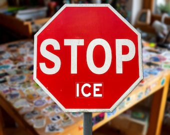 Può includere: Un cartello ottagonale in metallo rosso con bordo bianco e la parola STOP in grandi lettere maiuscole bianche. Sotto la parola STOP, la parola ICE è stampata in lettere maiuscole bianche più piccole. Il cartello è su un palo grigio.