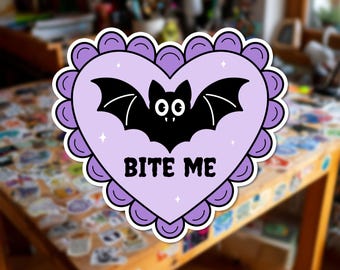 Può includere: Un adesivo a forma di cuore con bordo smerlato viola, raffigurante un pipistrello nero con occhi e zanne bianche, e la scritta BITE ME in lettere nere su sfondo viola chiaro, decorato con piccole stelle bianche a quattro punte.