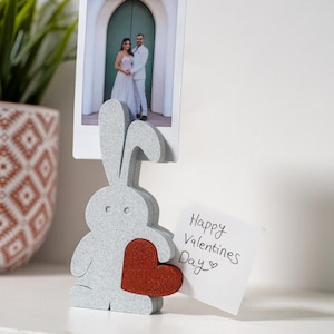 Puede incluir: Un portafotos gris con forma de conejo con un corazón rojo, que sostiene una foto de una pareja. Una pequeña nota dice "Happy Valentines Day". El soporte está sobre una superficie blanca, con una planta en maceta en el fondo.