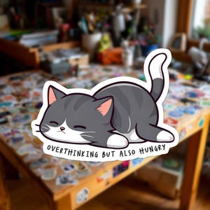 May include: A sticker depicting a cartoon cat in grey and white, lying down with closed eyes. The text "OVERTHINKING BUT ALSO HUNGRY" is below the cat. The cat has pink inner ears and white paws. The sticker is on a wooden table.