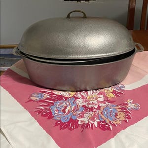 May include: A large, silver-colored cooking pot with a domed lid and a brass handle. The pot sits on a pink and white floral patterned tablecloth. The pot has two side handles.