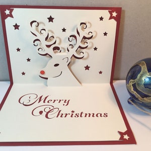 Op de afbeelding: Een witte pop-upkaart met een rode rand en een 3D-rendierontwerp. De kaart heeft de tekst "Merry Christmas" in rode cursieve letters. Het rendier heeft een rode neus en gekrulde geweien. De kaart is versierd met kleine rode sterren.
