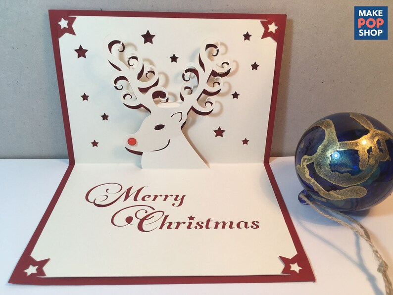 Op de afbeelding: Een witte pop-upkaart met een rode rand en een 3D-rendierontwerp. De kaart heeft de tekst "Merry Christmas" in rode cursieve letters. Het rendier heeft een rode neus en gekrulde geweien. De kaart is versierd met kleine rode sterren.