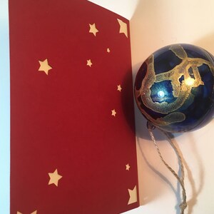 Op de afbeelding: Een rode kaart met uitgesneden sterren. De kaart is dubbelgevouwen en de bovenste helft is zichtbaar. Een blauwe glazen kerstbal met gouden verf hangt aan een touwtje.