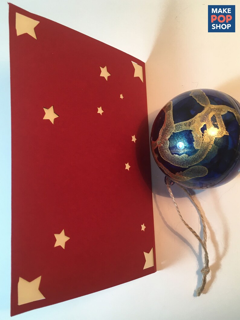 Op de afbeelding: Een rode kaart met uitgesneden sterren. De kaart is dubbelgevouwen en de bovenste helft is zichtbaar. Een blauwe glazen kerstbal met gouden verf hangt aan een touwtje.