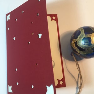 Op de afbeelding: Een rode kaart met witte sterren die eruit zijn geknipt. De kaart is doormidden gevouwen en de binnenkant is cr&egrave;mekleurig. Op de achtergrond is een blauw en gouden ornament met een touwtje te zien.