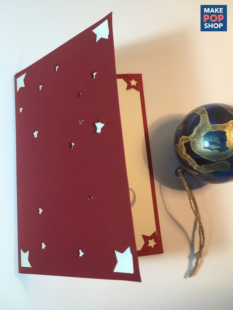 Op de afbeelding: Een rode kaart met witte sterren die eruit zijn geknipt. De kaart is doormidden gevouwen en de binnenkant is cr&egrave;mekleurig. Op de achtergrond is een blauw en gouden ornament met een touwtje te zien.