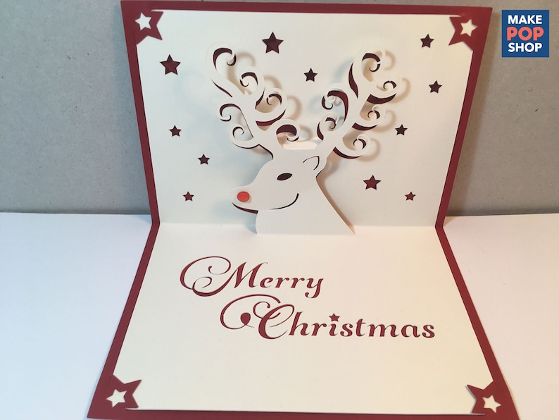 Op de afbeelding: Een witte papieren pop-upkaart met een rode rand. De kaart heeft een 3D-rendier met een rode neus en gekrulde geweien. De kaart heeft de tekst "Merry Christmas" in rode cursieve letters.