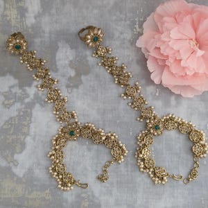 May include: Two gold-toned hand harnesses featuring intricate filigree metalwork, small white beads, and central green stones. Each piece consists of a ring connected by a decorative chain to a wrist bracelet, showcasing a detailed floral-inspired design.