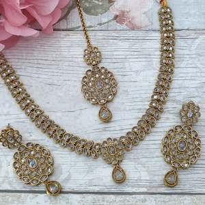 May include: A gold-coloured jewellery set comprising a necklace, earrings, and a headpiece. The set is decorated with clear, round gemstones and teardrop-shaped pendants. The necklace and earrings have a floral design. The headpiece has a central gemstone and a teardrop pendant.