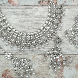 May include: A silver and white bridal jewellery set with a necklace, earrings, and a maang tikka. The necklace is made of silver with a mirrored finish and features a layered design with pearl accents. The earrings are also silver with a mirrored finish and feature a teardrop design with pearl accents. The maang tikka is a silver chain with a mirrored pendant and pearl accents.
