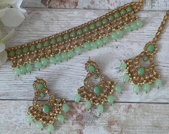 May include: A gold-toned jewelry set featuring a necklace, earrings, and a headpiece. The set is adorned with light green oval-shaped stones and small white beads. The necklace and headpiece have a similar design, with the earrings having a teardrop shape.