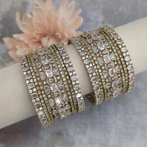 May include: A set of two gold-toned bangles featuring multiple rows of clear, faceted stones in various shapes, including rectangular, oval, and round cuts, arranged in a dense, intricate pattern that covers the entire surface of each bracelet.