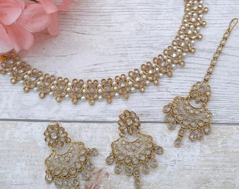 May include: A gold necklace, earrings, and a tikka with white pearl and clear rhinestone accents. The jewelry is on a white wooden surface.