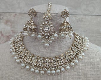 May include: A gold-toned bridal jewelry set featuring a necklace, earrings, and a headpiece. The set is adorned with clear crystals and faux pearls. The necklace and earrings have a bell shape. The headpiece is a central pendant with a chain.