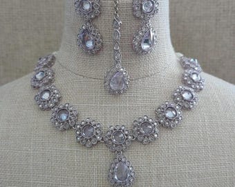 Könnte beinhalten: Ein silberfarbenes Schmuckset mit Halskette, Ohrringen und Kopfschmuck. Die Halskette und die Ohrringe haben ein Blumenmuster mit klaren, runden und tropfenförmigen Steinen. Der Kopfschmuck hat einen tropfenförmigen Stein.