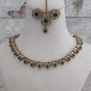 Op de afbeelding: Een goudkleurige sieradenset met een ketting, oorbellen en een hoofdband. De ketting en oorbellen hebben een herhalend patroon van heldere en donkergroene stenen. De hoofdband heeft een centrale hanger met een bijpassende groene steen.