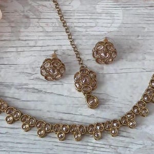 May include: A gold necklace with a delicate chain and clear stones, a matching pair of earrings, and a matching maang tikka.