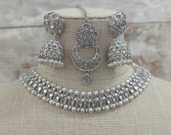 May include: A silver-toned jewelry set featuring a necklace, earrings, and a headpiece. The necklace and earrings have a combination of clear stones and small pearls. The headpiece has a crescent moon design with a central stone.