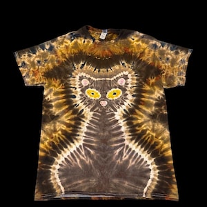 May include: Brown and tan tie-dye t-shirt with a cat face design. The cat's face has yellow eyes and pink accents. The shirt is a short-sleeved crew neck style, suitable for casual wear.