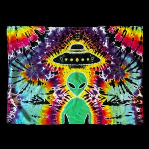 Może przedstawiać: Tapiseria tie-dye przedstawiająca zielonego kosmitę z czarnym UFO nad nim. Tapiseria ma psychodeliczną kolorystykę z odcieniami żółtego, pomarańczowego, czerwonego, różowego, niebieskiego, zielonego i czarnego.