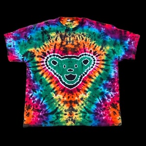 May include: A tie-dye t-shirt with a vibrant array of colors, including green, orange, pink, and blue. The shirt features a green bear graphic with a white outline. The shirt is a short-sleeved crew neck.
