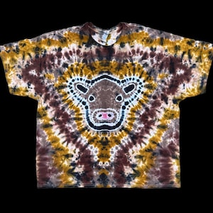 May include: A tie-dye t-shirt with a brown cow face design in the center. The shirt is a blend of yellow, brown, and black colors.