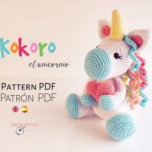 Puede incluir: Un juguete de amigurumi de unicornio tejido a crochet con un corazón rosa en sus brazos. El unicornio es blanco con una cara azul claro y un cuerno amarillo. La melena y la cola son de colores arcoíris.