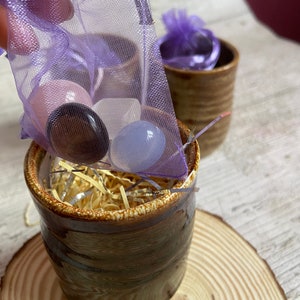 May include: A small brown ceramic pot filled with colourful stones and shredded paper. The pot is sitting on a wooden coaster. A purple mesh bag with more stones is being poured into the pot.