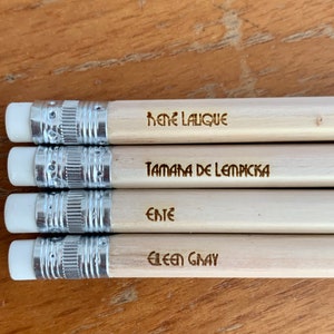 Puede incluir: Cuatro lápices con gomas blancas y bandas metálicas plateadas. Cada lápiz es de color claro con texto: "René Lalique", "Tamara de Lempicka", "Ente" y "Eileen Gray". Los lápices están colocados sobre una superficie de madera.
