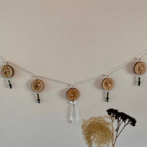 Puede incluir: Una guirnalda de madera con fases lunares con cuentas negras que cuelgan de cada luna. La guirnalda está enhebrada en un alambre transparente y tiene un cristal blanco colgando de la luna del centro.