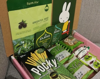 May include: A pink and white cardboard box filled with green tea flavored snacks. The box contains a package of Pocky green tea sticks, a package of matcha green tea cookies, and several individual packages of matcha flavored snacks. The box also includes a green tea face mask and a small green tea bag.