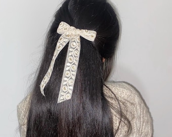 May include: A white hair bow with gold floral embroidery. The bow is tied in a simple knot and is worn in the hair of a person with long black hair.