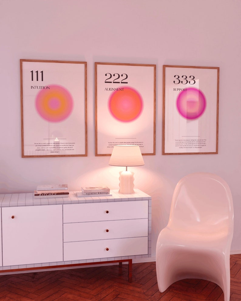 May include: Three framed prints with pink and orange gradient circles and the numbers 111, 222, and 333 with the words "Intuition", "Alignment", and "Support" below each number. The prints are hanging on a white wall above a white dresser with three drawers and a white chair.