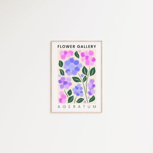 May include: A framed print with a light beige background featuring a stylized illustration of purple and pink flowers with green leaves. The text "FLOWER GALLERY" and "AGERATUM" is printed in black at the top and bottom of the print.