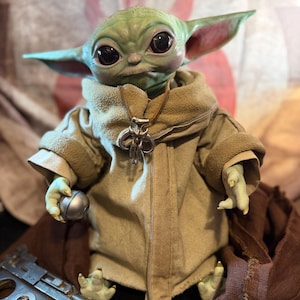 May include: A detailed figurine of a green-skinned character with large black eyes and pointed ears, wearing a tan robe and a silver pendant. The figure holds a silver ball and is posed with a blaster pistol.