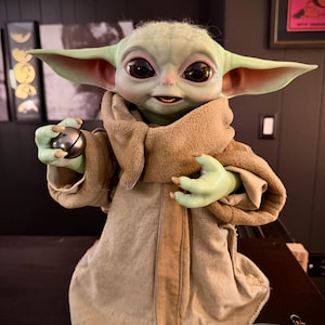 Puede incluir: Figura detallada de un personaje de una popular franquicia de ciencia ficción. La figura tiene ojos grandes, piel verde y orejas grandes. Lleva una túnica marrón y sostiene una esfera plateada. La figura está sobre una superficie oscura.