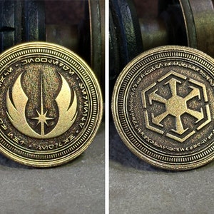 May include: Two gold-coloured metal coins with different symbols. The first coin features a stylised symbol of a lightsaber and wings with the text "The Force Will Be With You, Always" around the edge. The second coin features a stylised symbol of a cross with the text "The Empire Strikes Back" around the edge.