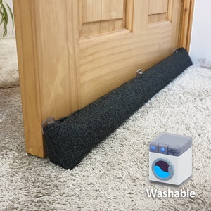 Può includere: Un fermaporta nero per le correnti d'aria con un rivestimento in tessuto grigio. Il fermaporta è posizionato sotto una porta in legno e si trova su un tappeto bianco. Il testo "Lavabile" è sotto un'icona di una lavatrice.