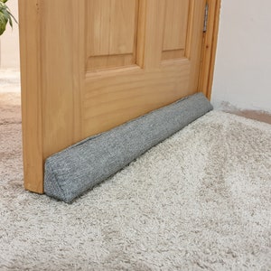 Può includere: Un fermaporta grigio in tessuto è posizionato nella parte inferiore di una porta in legno. La porta è aperta e il fermaporta è su un tappeto grigio chiaro.