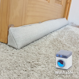 Peut inclure: Un butoir de porte gris avec un revêtement en tissu et une forme triangulaire. Le butoir de porte est placé sur un tapis gris clair. Le butoir de porte est lavable, comme indiqué par l'icône de la machine à laver.