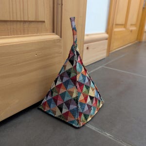 May include: A colourful triangular doorstop with a geometric pattern. The doorstop is made of fabric and has a loop handle.