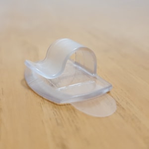 Könnte beinhalten: Ein transparenter Kabelclip aus Kunststoff zum Organisieren von Kabeln und Drähten. Der Clip hat ein flexibles Design, das es ermöglicht, ihn auf verschiedenen Oberflächen zu verwenden.