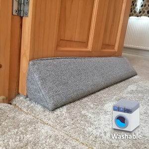 Può includere: Un fermaporta a cuneo in tessuto grigio posizionato su un tappeto grigio chiaro. Il fermaporta ha la forma di un cuneo e una parte superiore piatta. Il fermaporta è lavabile, come indicato dall'icona della lavatrice con il testo "Washable" sotto.