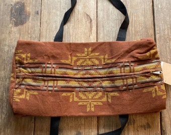 May include: A brown canvas tote bag with a geometric pattern in shades of yellow and brown. The bag has black straps and a zipper closure.