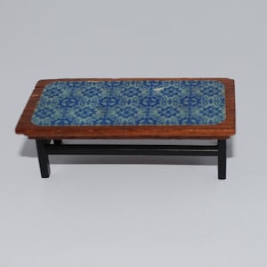 May include: A miniature wooden coffee table with a dark brown frame and black legs. The tabletop features a blue and white patterned design. This small table is suitable for a dollhouse or miniature setting.