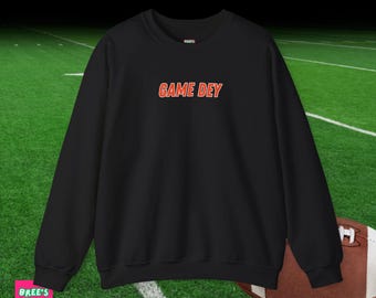 May include: Black crew neck sweatshirt with the text "GAME DEY" in orange lettering. The sweatshirt is displayed on a green field with a football in the background. The brand "Bree's Tees" is in the lower left corner.
