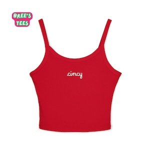 Puede incluir: Un top de tirantes rojo con tirantes finos. La palabra "cincy" está escrita en escritura blanca en la parte delantera. La parte superior tiene un escote redondo. La esquina superior izquierda tiene un logotipo rosa y verde que dice "Bree's Tees".